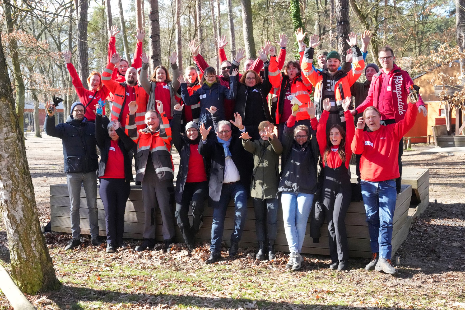 Eine Gruppe von Menschen posiert jubelnd in einem Wald. Viele tragen orangerote Arbeitskleidung.