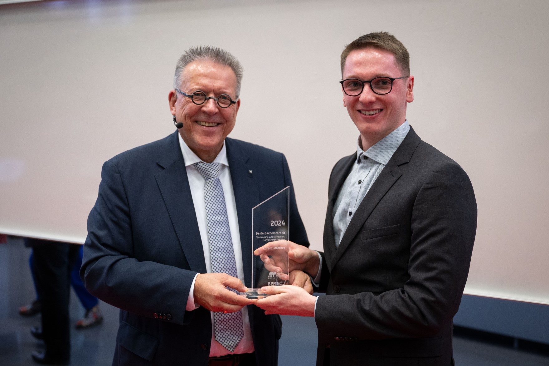 Two men in suits hold an award in their hands and smile into the camera.