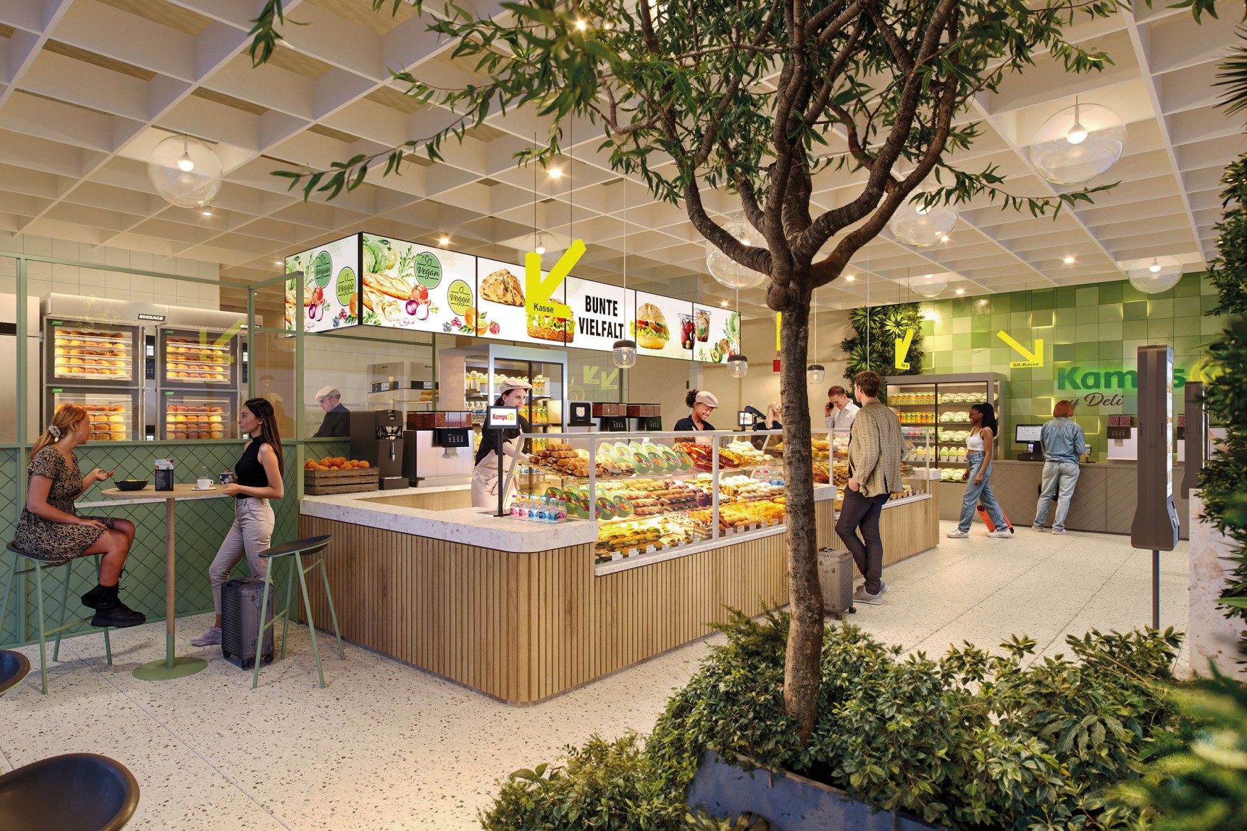Kamps my Deli Geschäft von Innen, ein kleiner Baum im Vordergrund, im Hintergrund ein Tresen, bestückt mit Essen, Personen links und rechts vom Tresen