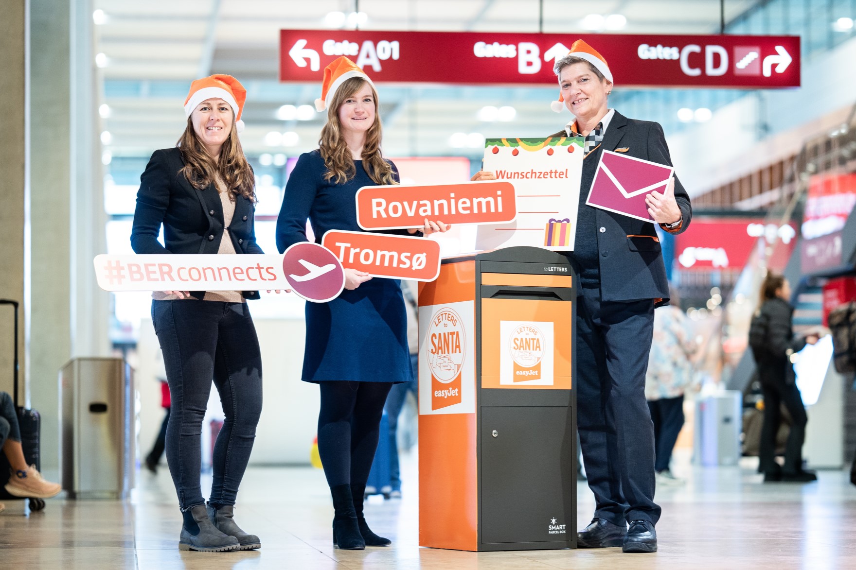 Drei Personen stehen mit Nikolausmütze im Terminal und halten Schilder hoch. Zwischen den Personen steht ein großer Briefkasten.