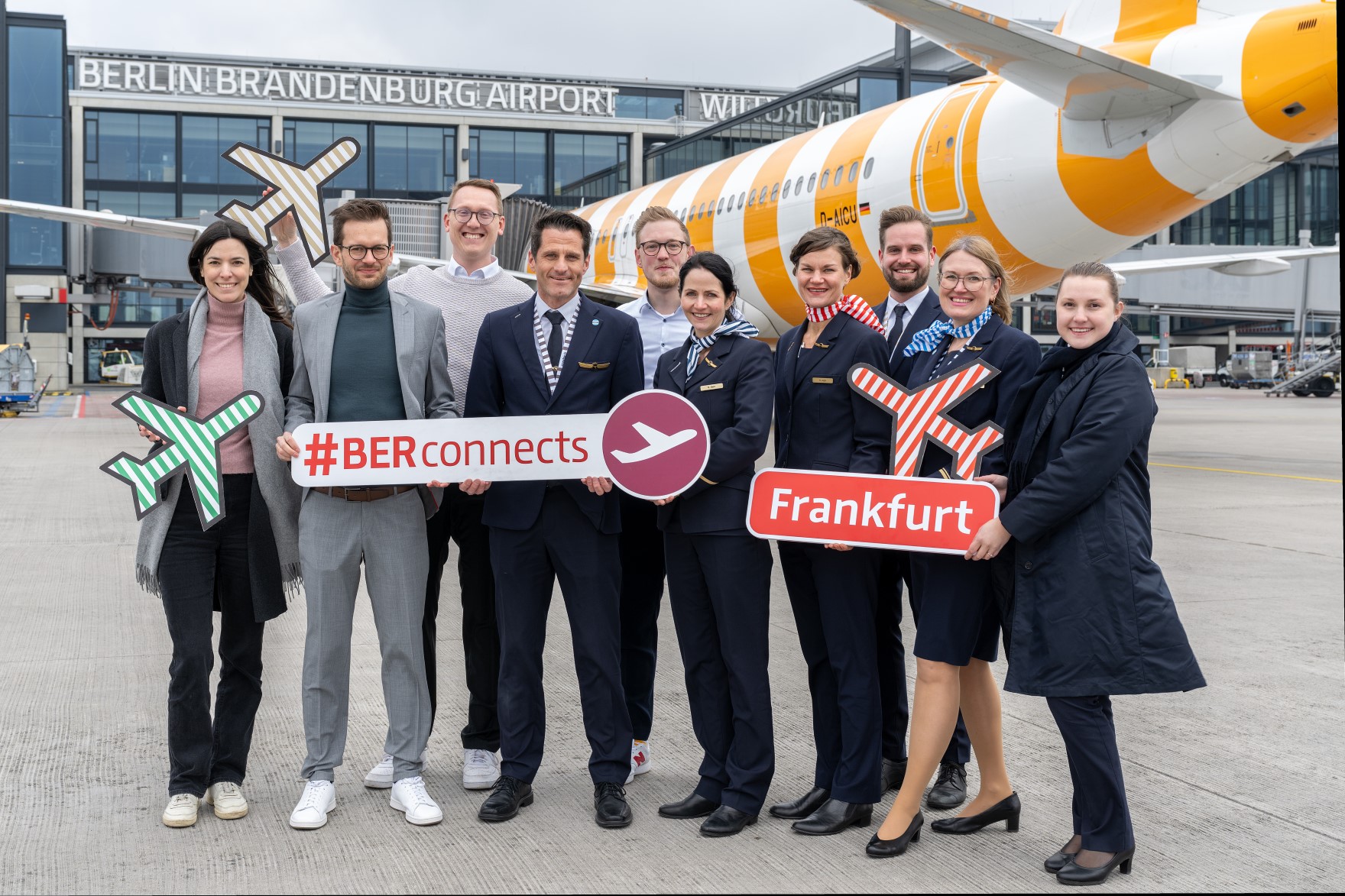 Eine Gruppe von zehn Personen, die einen in Business-Outfits, die anderen in den Uniformen einer Flugzeug-Crew posiert auf dem Vorfeld des BER vor einem gelb-weiß gestreiften Flugzeug. Die Personen halten Schilder mit Flugzeug-Piktogrammen und den Aufschriften "#BERconnects" und "Frankfurt". / English: A group of ten people, some in business outfits, others in the uniforms of an aircraft crew, pose on the apron of BER in front of a yellow and white striped aircraft. The people are holding signs with aircraft pictograms and the words ‘#BERconnects’ and ‘Frankfurt’.