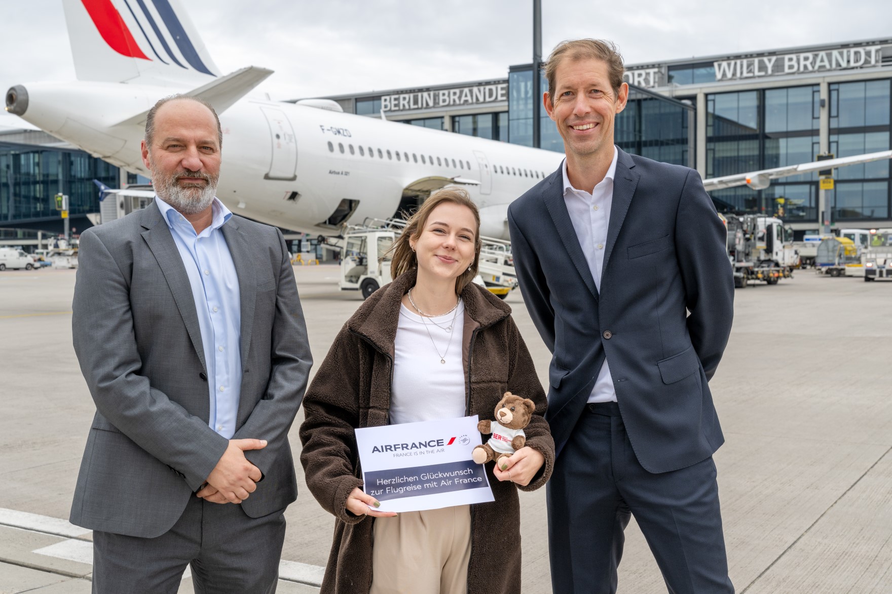 Zwei Männer und eine Frau posieren auf dem Vorfeld vor einem Flugzeug. Die Frau, in der Mitte, präsentiert einen Gutschein und hält einen Teddy im Arm.