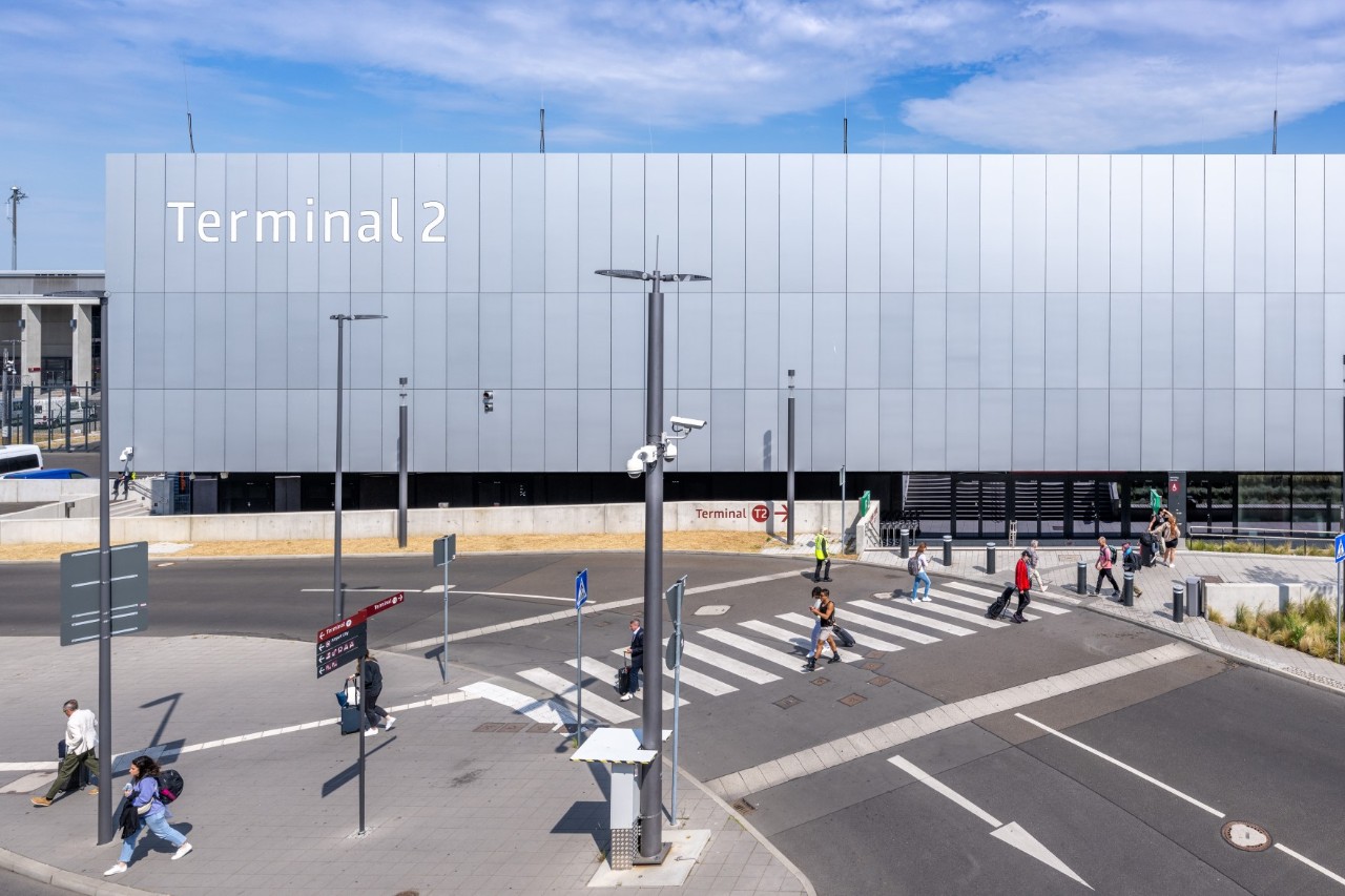 Das Terminal 2 von außen. Davor eine Straße und Bäume. Menschen passieren die Straße.