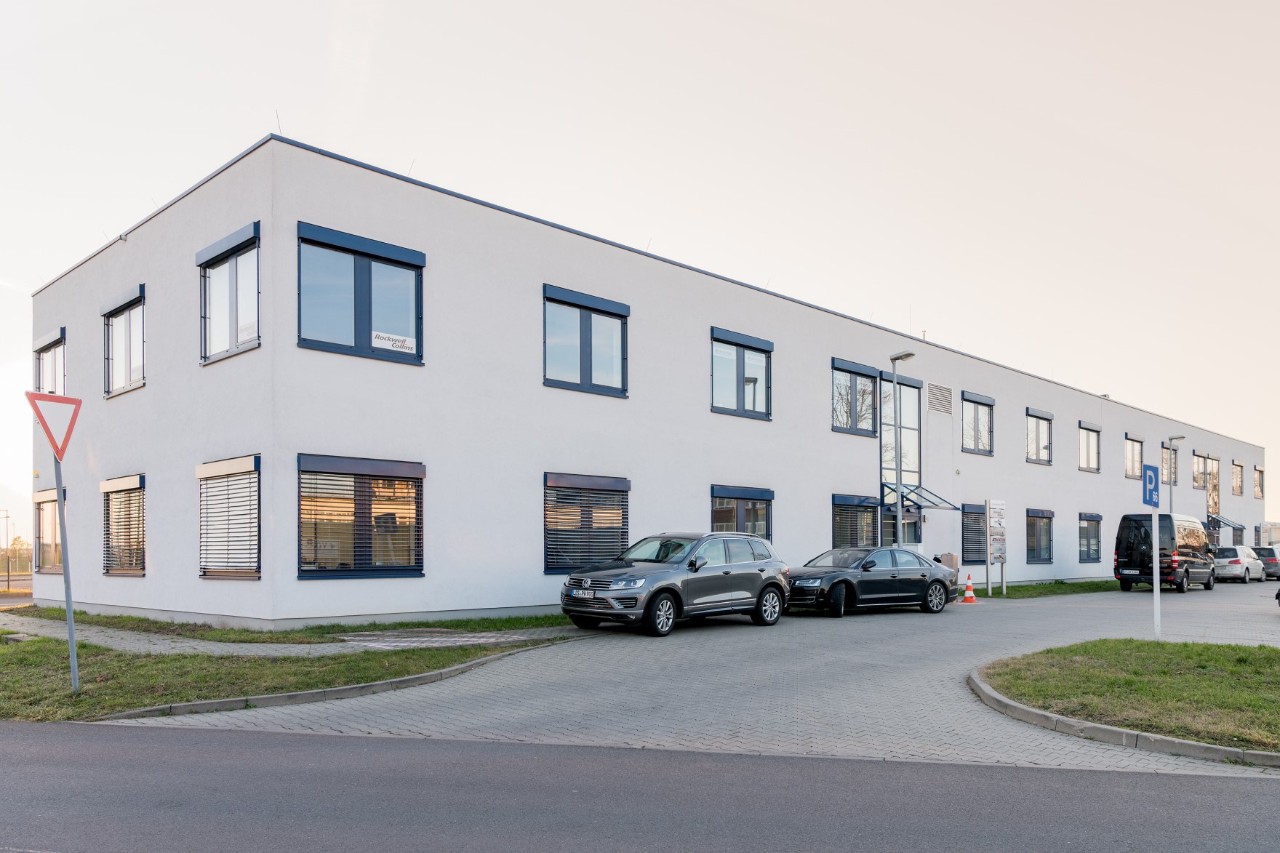 Ein weißes zweistöckiges Gebäude.. Davor Autos und eine Straße.