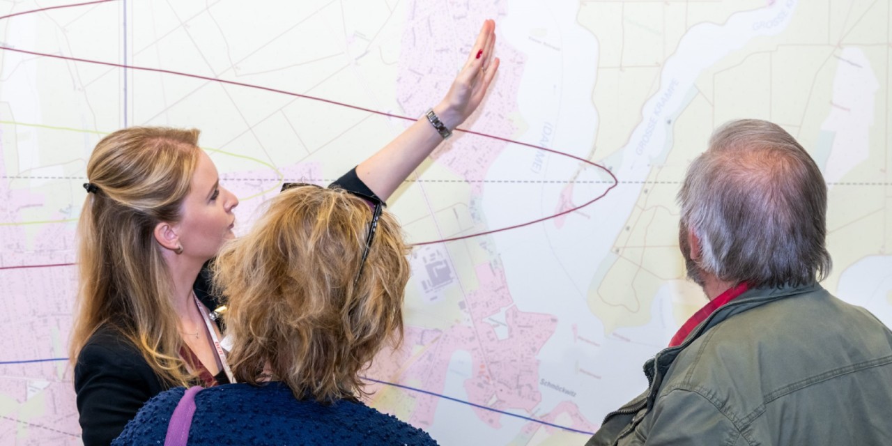 Drei Personen stehen vor einer großen Wandkarte, eine von ihnen erklärt und zeigt etwas auf der Karte.