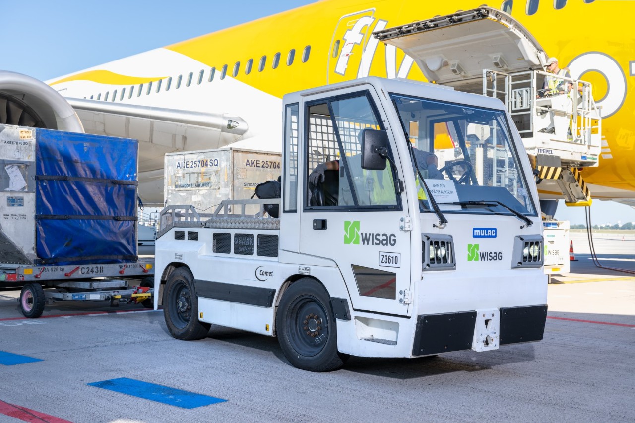 Ein Gepäckwagenschleppfahrzeug und Gepäckcontainer stehen neben einem Flugzeug, das gerade be- oder entladen wird.