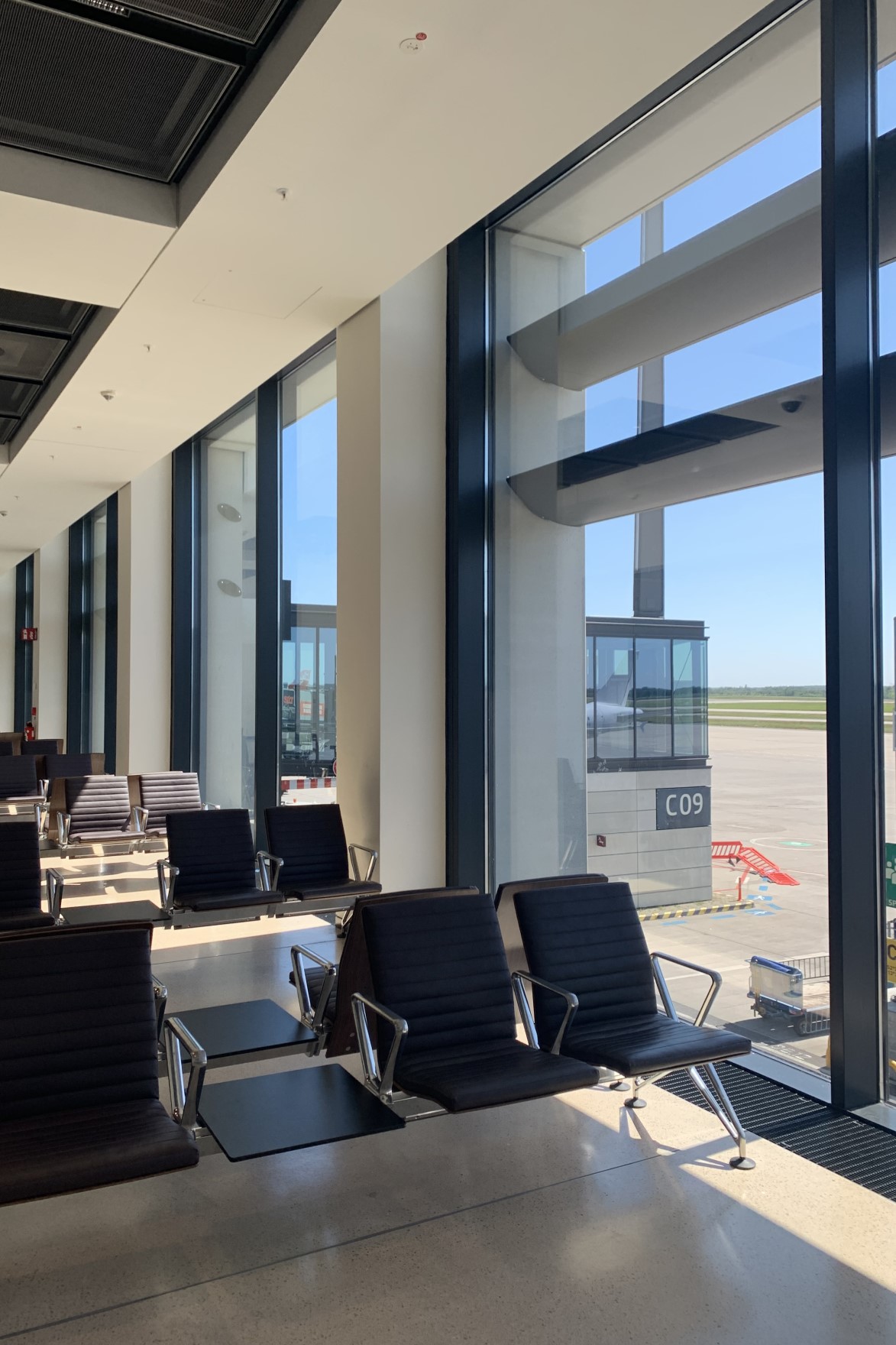 Sitzbereich im Flughafenterminal. Große Fenster, an denen oben Verschattungsmodule angebracht sind, ermöglichen den Blick auf das Vorfeld.