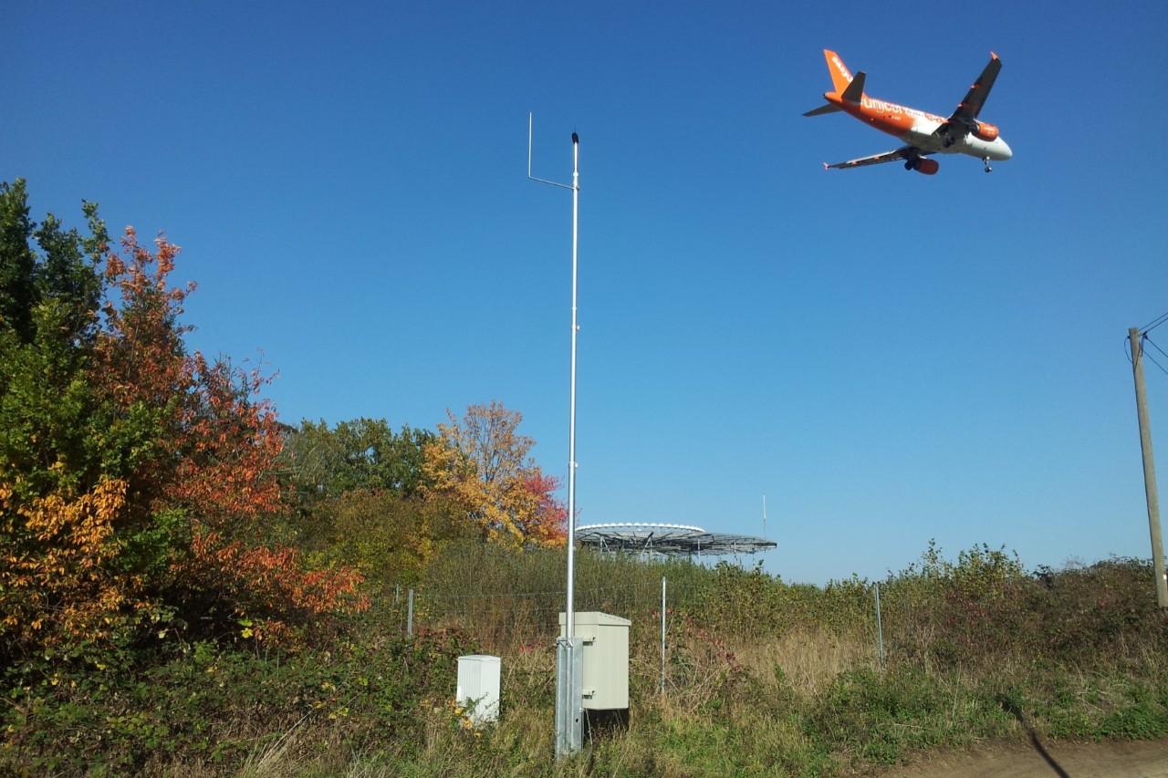 Ein Flugzeug fliegt über eine stationäre Fluglärmmessstelle.