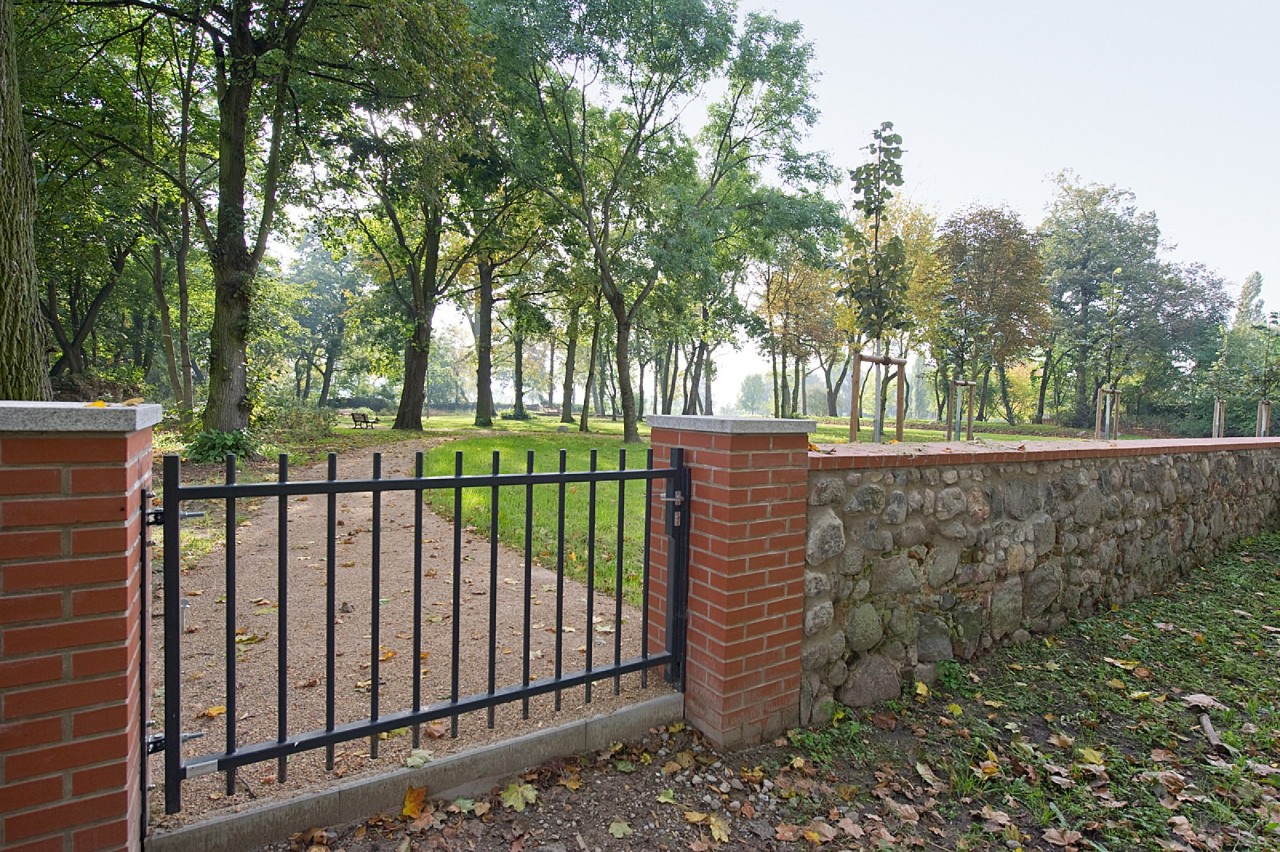 Eine gepflegte Parkanlage mit Bäumen und einem Weg ist von einer Mauer, die abschnittsweise in einen Zaun übergeht, umgeben.