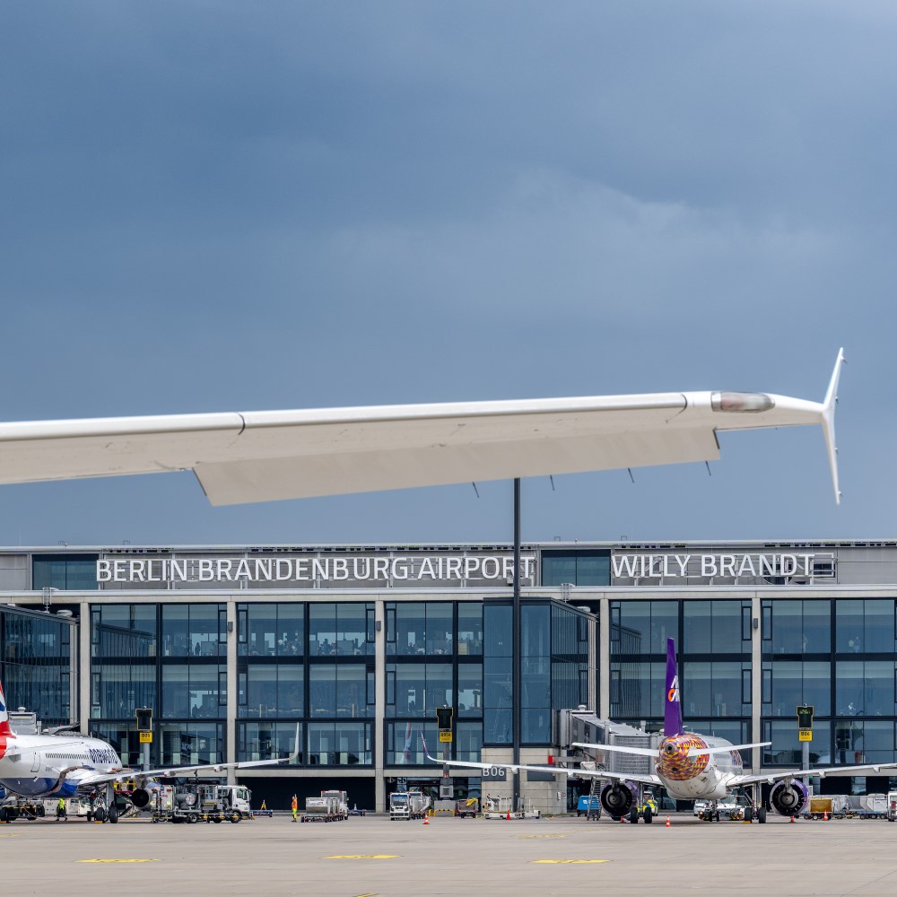 Im Vordergrund ein weißer Flugzeug-Flügel, dahinter 2 Flugzeuge am Terminal 1.