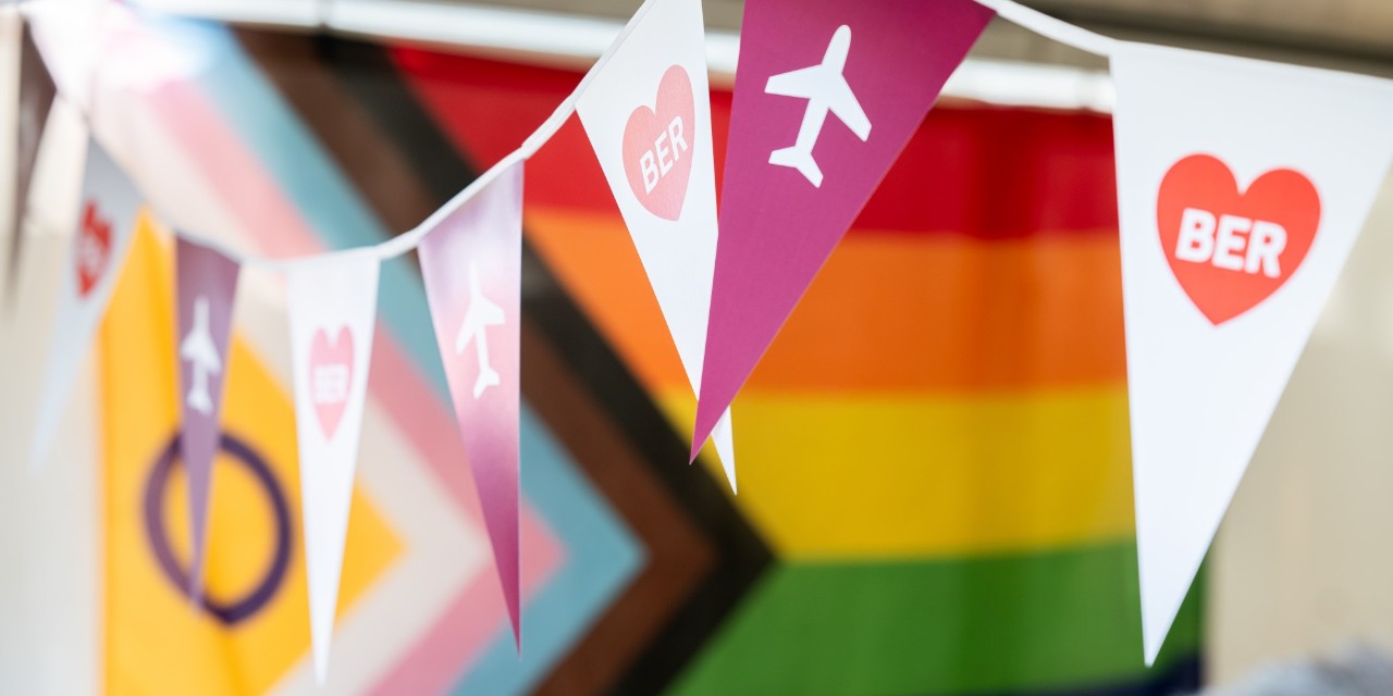Eine Girlande mit BER-Piktogrammen hängt neben einer Regenbogenflagge.