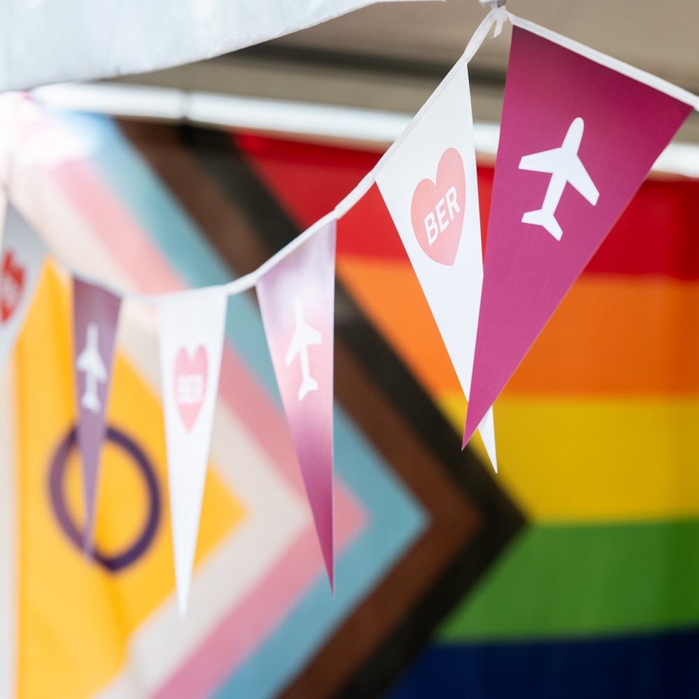 Eine Girlande mit BER-Piktogrammen hängt neben einer Regenbogenflagge.
