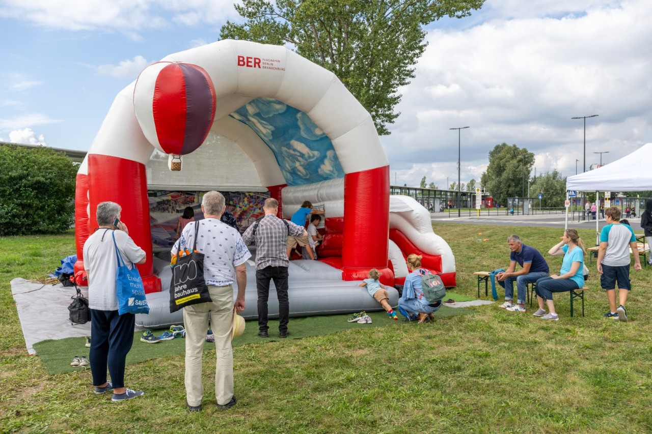 Eine Hüpfburg mit dem Logo des BER ist auf einer Wiese aufgestellt. Kinder hüpfen darauf, Erwachsene stehen darum herum.