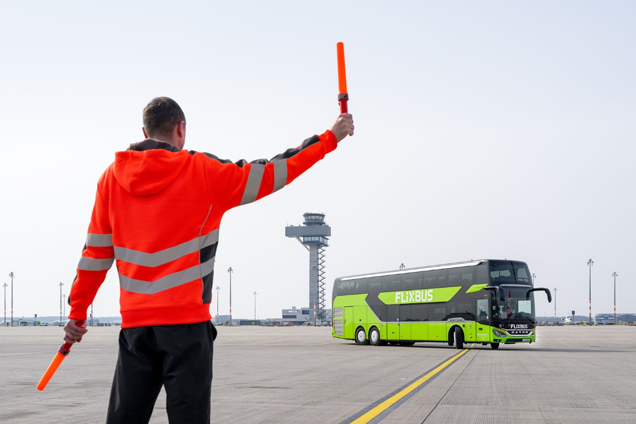 Ein Mann steht mit Warnweste auf dem Vorfeld. Im Hintergrund fährt ein grüner Fernbus.