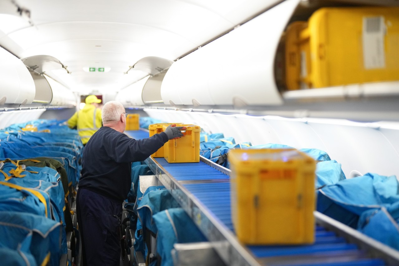 Im Passagierraum eines Flugzeugs sind die Sitze abgedeckt und über die Lehnen einer Sitzreihe ist ein Laufband gelegt, über das Kisten in den hinteren Bereich des Flugzeugs transportiert werden.