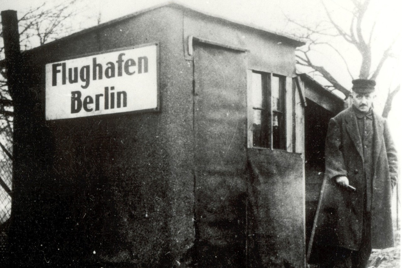 Eine zerbrechliches Gebäude mit dem Schriftzug "Flughafen Berlin". Davor ein Mensch. Das Bild ist schwarz-weiß.