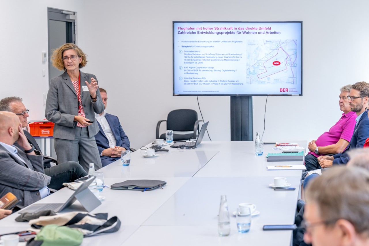Personen in Businesskleidung sitzen um einen Konferenztisch herum. Eine Person steht seitlich am Tisch und referiert. Am Kopfende des Tisches steht ein großer Bildschirm, auf dem eine Präsentation gezeigt wird.