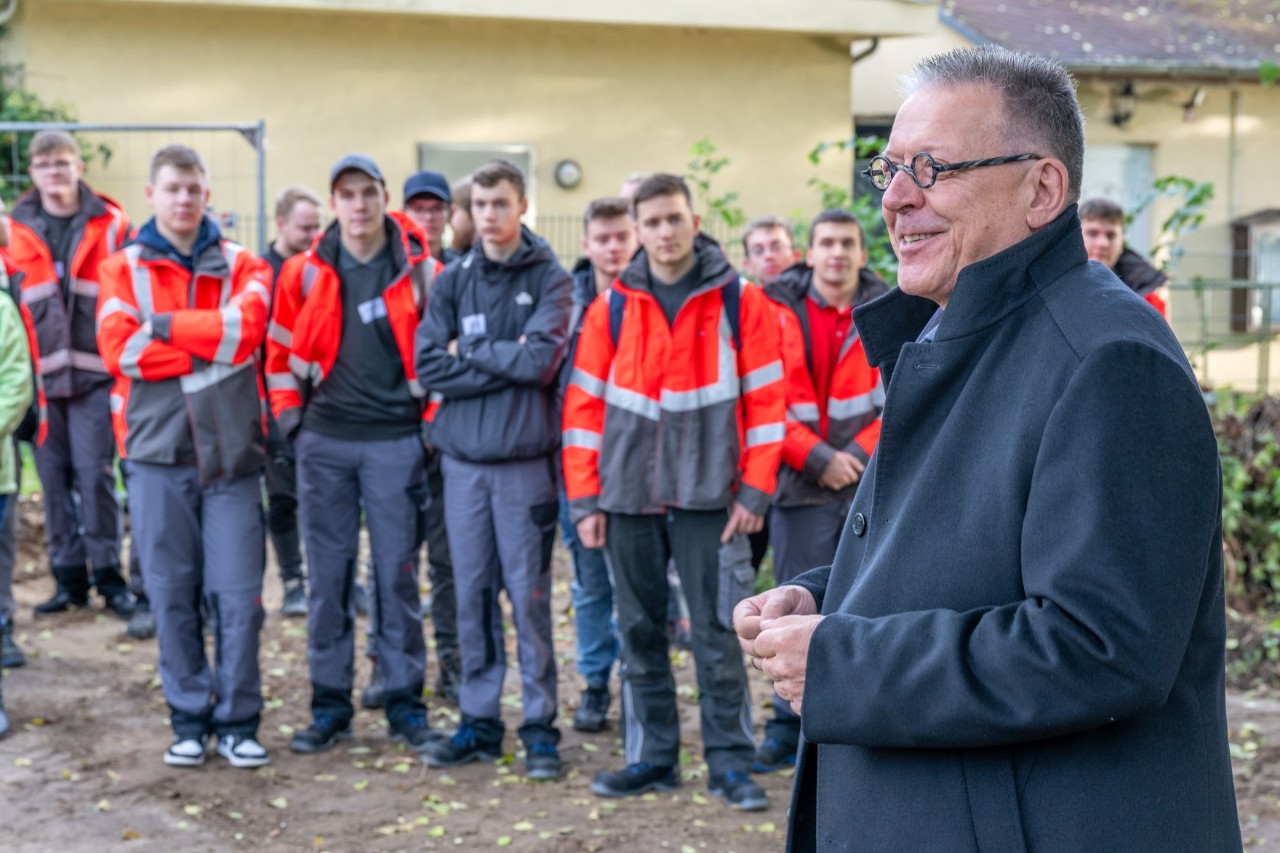 Ein Mann im Vordergrund hält eine Rede. Hinter ihm stehen Personen in Warnjacken und hören zu.