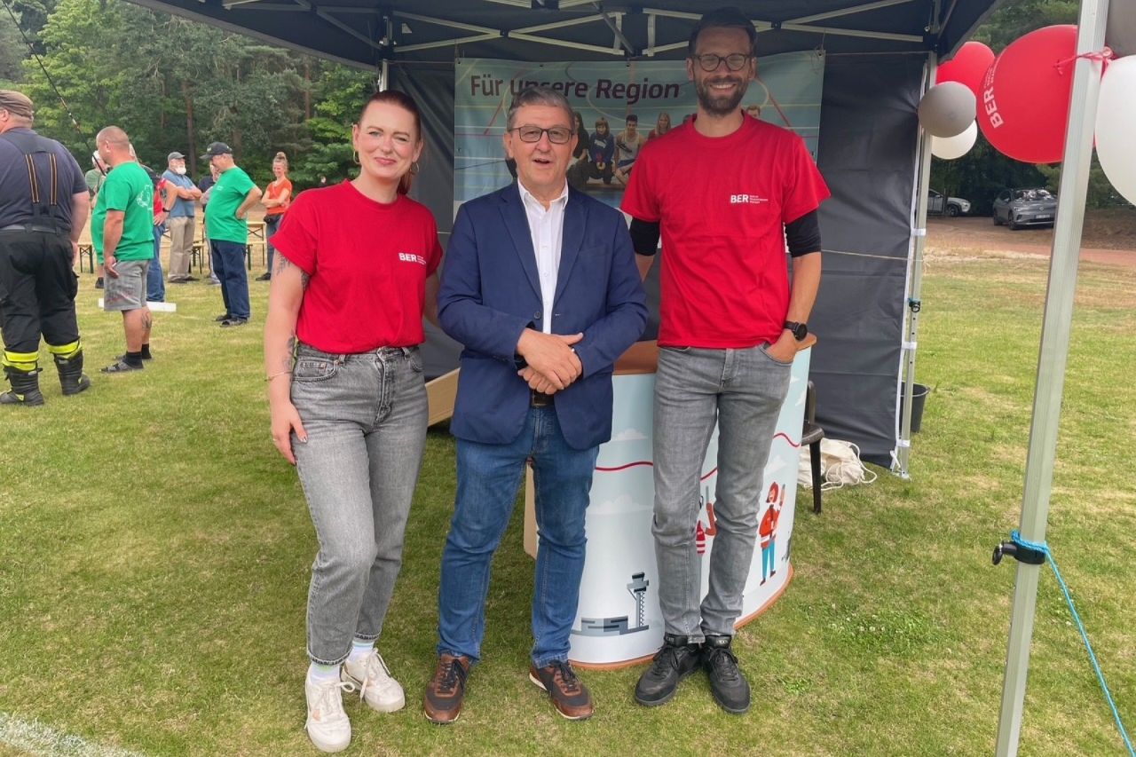 Drei Personen stehen an einem BER-Infostand. Im Hintergrund sind mehrere Besucher zu sehen.