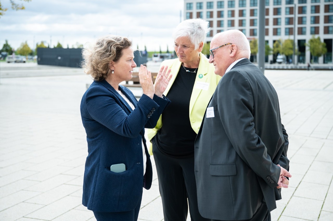 Drei Personen unterhalten sich draußen auf einem großen Platz.