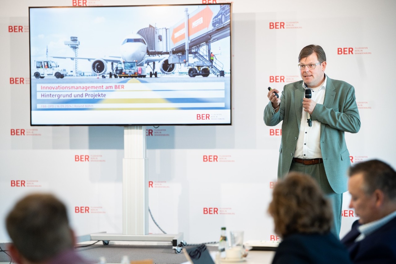 Links steht ein größerer Monitor. Rechts daneben hält eine Person einen Vortrag.