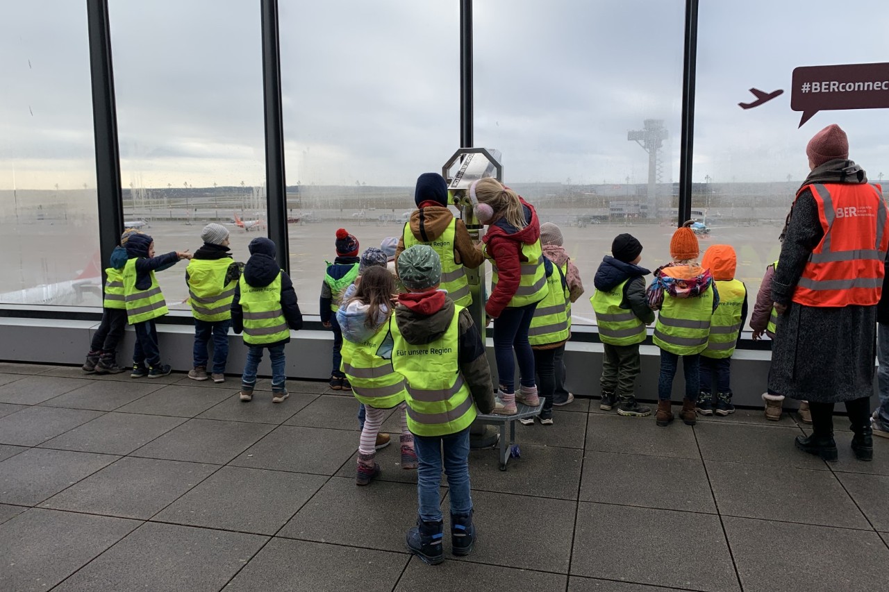 Kinder in gelben Warnwesten stehen vor einer großen Glasfront, von der aus man das Vorfeld eines Flughafens sieht.
