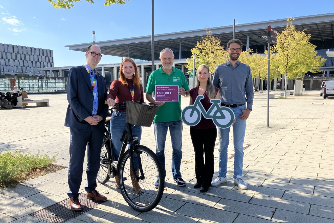 Fünf Menschen posieren lächelnd auf dem Platz vor Terminal 1 des BER. Eine Person hält ein Fahrrad, zwei andere Schilder.