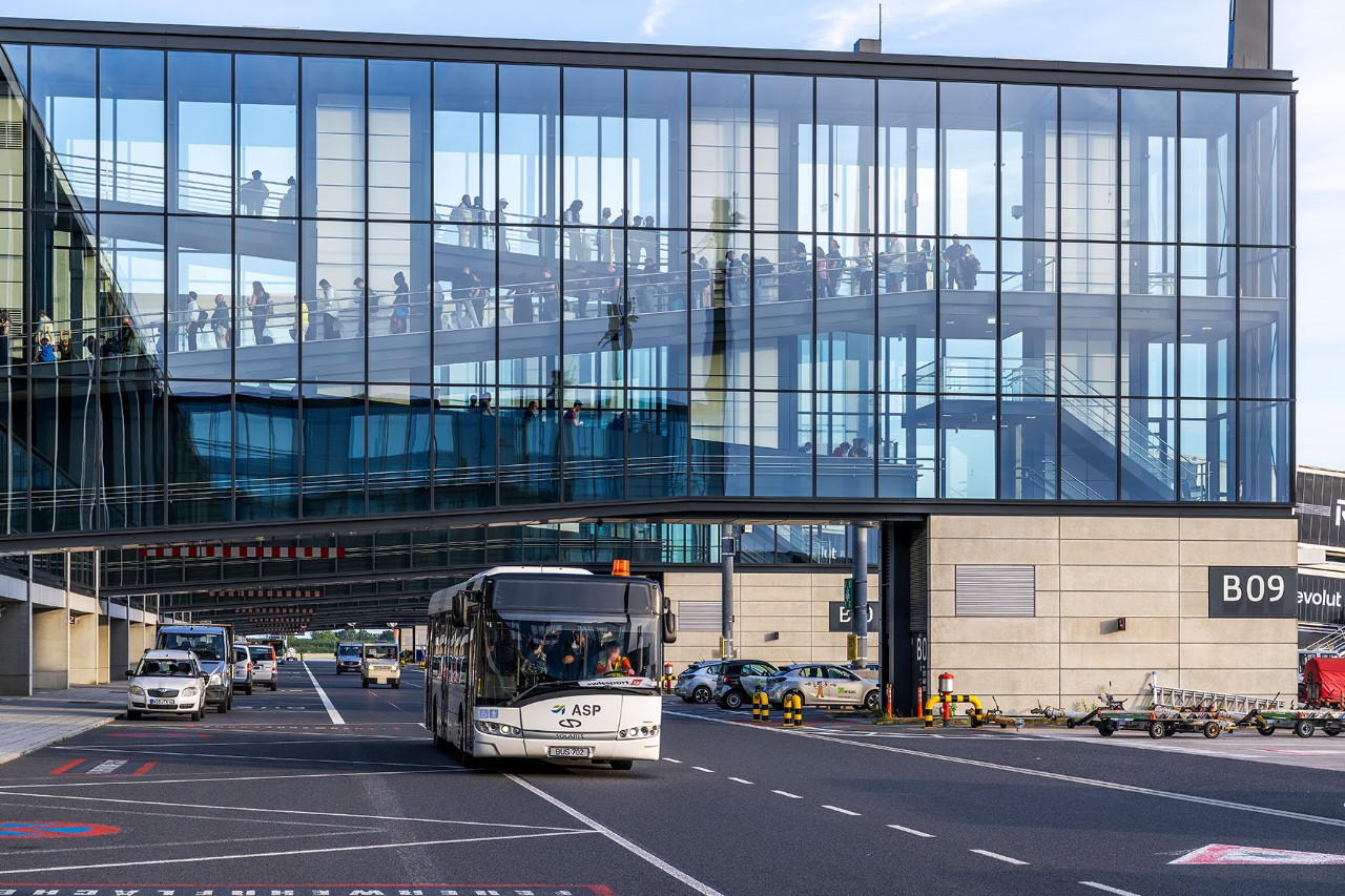 Ein Bus fährt auf der Straße im Sicherheitsbereich. Im Hintergrund ist eine Fluggastbrücke zu sehen.