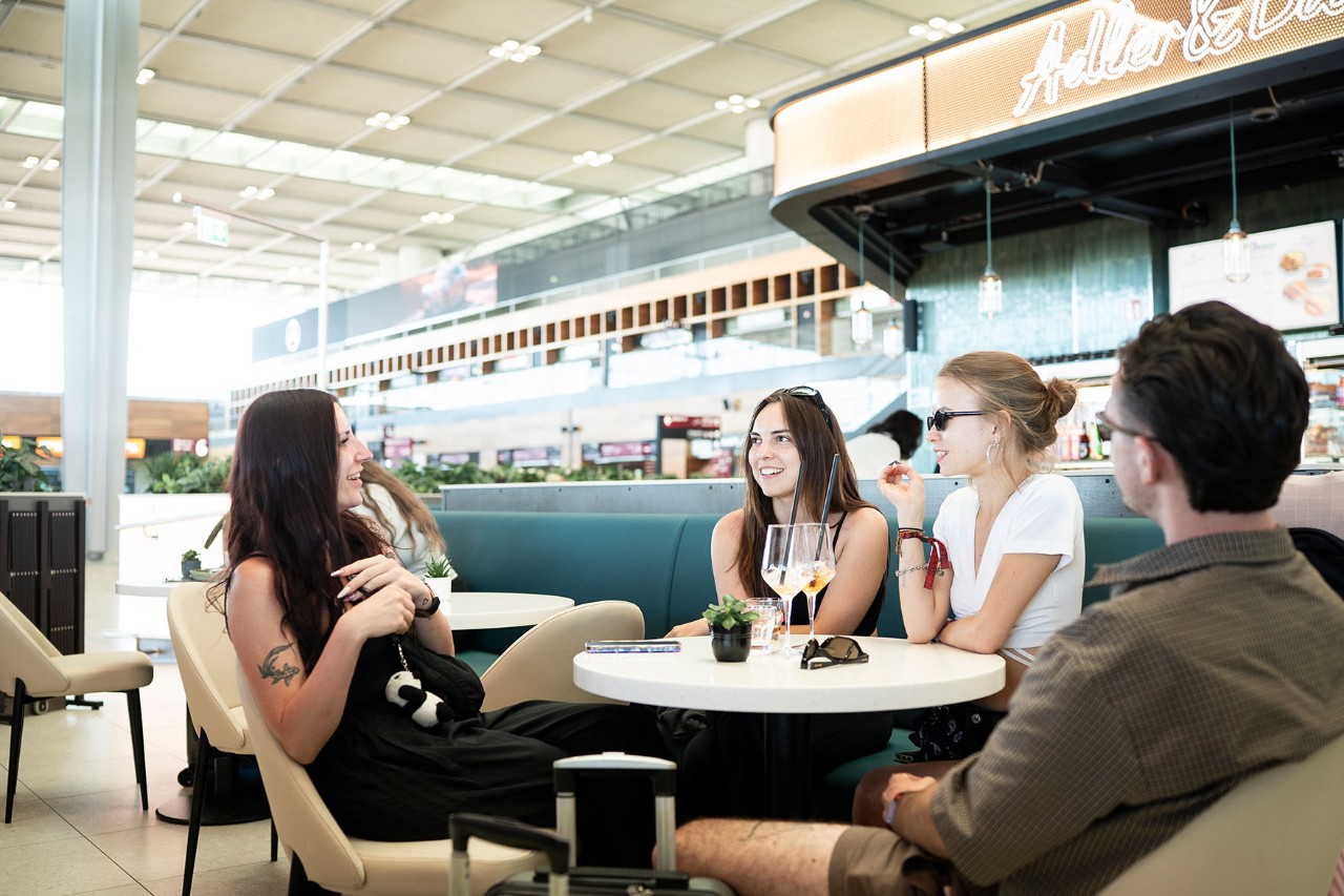 Eine junge Reisegruppe sind fröhlich in einer Bar am BER. Im Hintergrund ist das Terminal zu sehen.