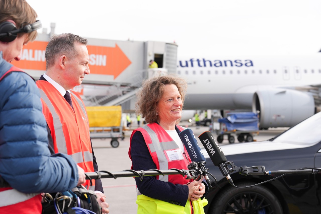 Ein Mann und eine Frau stehen vor einem Flugzeug der Airline Lufthansa und geben ein Interview