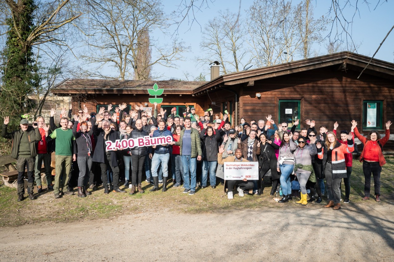 A large group of people pose smiling and cheering in front of a flat wooden house in a rural setting. Various signs are held up. One of them reads "2,400 trees".