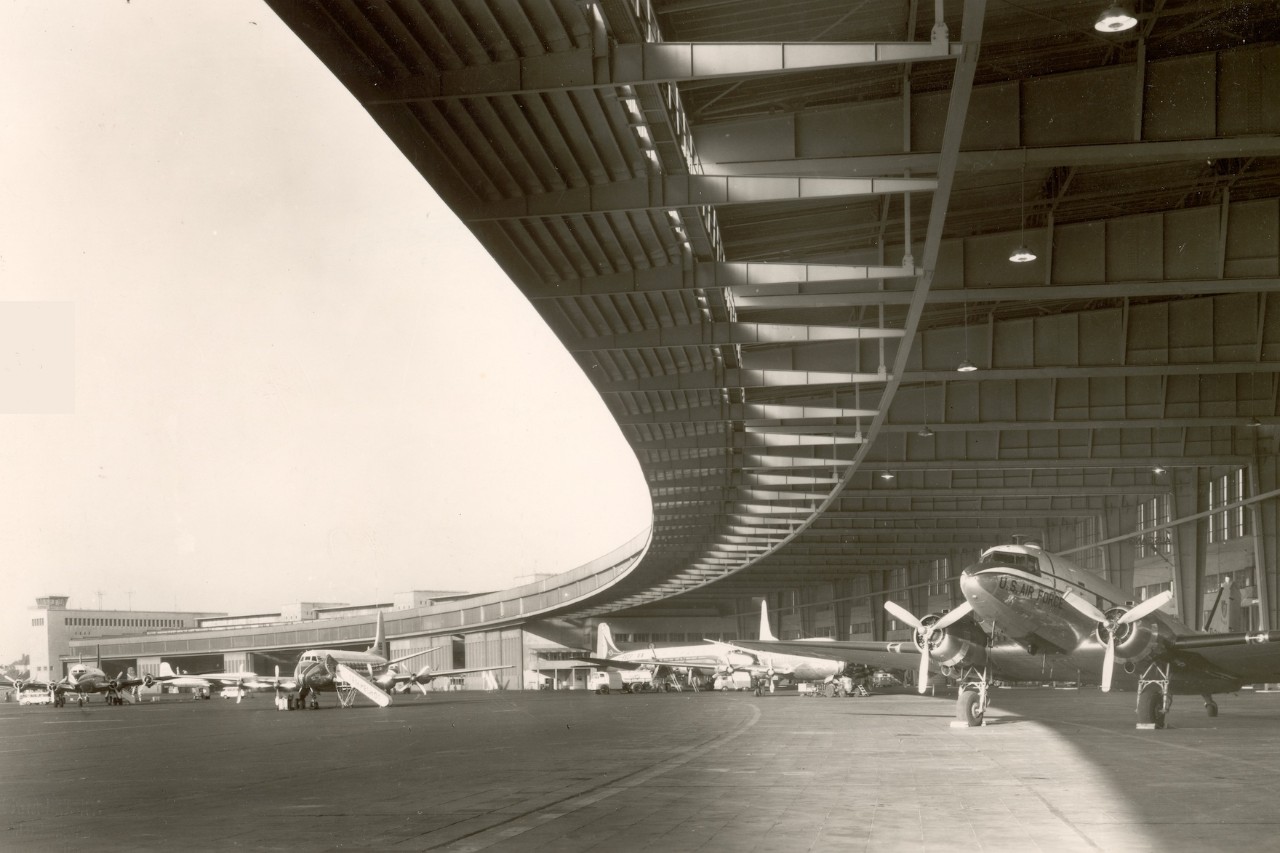 A black and white picture. Several propeller-driven aircraft are parked on the roofed apron.