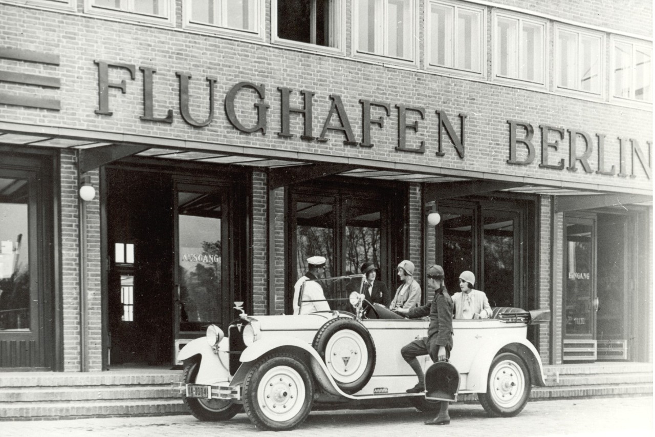 A black and white picture. An old car is parked in front of the building with the lettering "Berlin Airport". Several people are sitting in it.