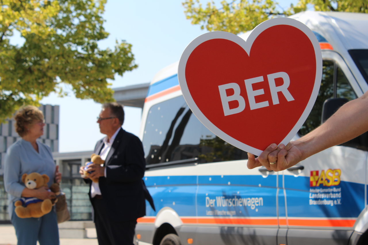 Two people are talking in the left background. In the right foreground, someone is holding up a heart-shaped sign with the word ‘BER’ on it.