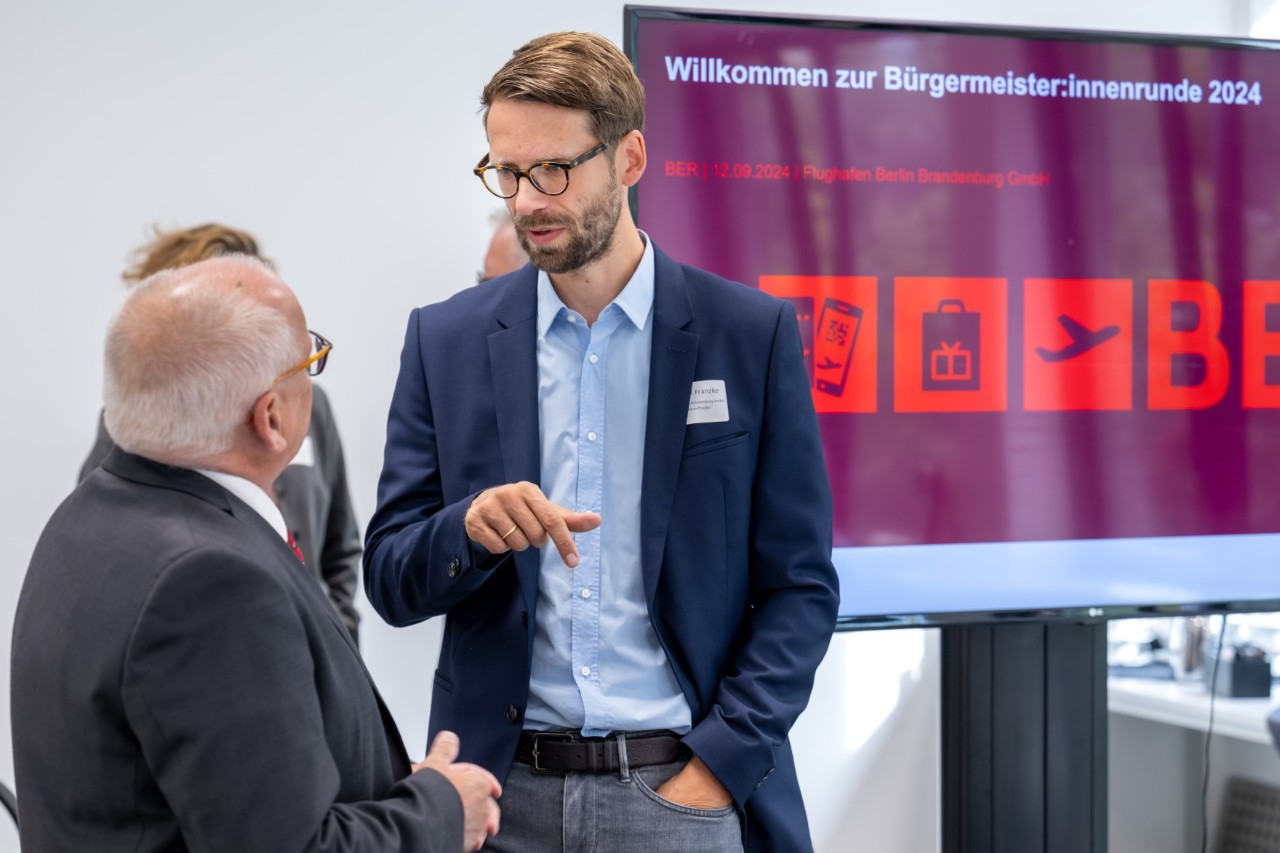 Two people in suits in conversation. In the background is a screen showing a presentation entitled "Willkommen zur Bürgermeister:innenrunde 2024" (‘Welcome to the 2024 mayors’ round’).