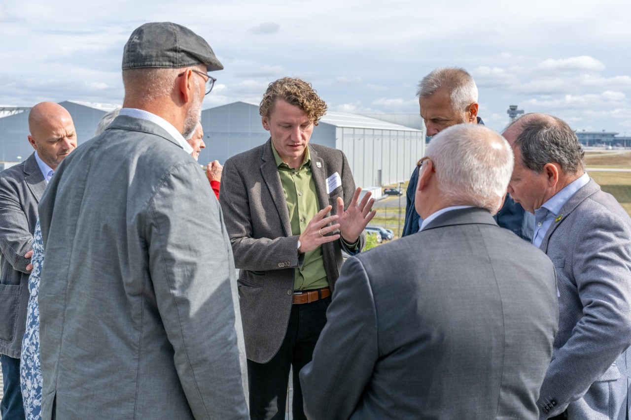 A group of people in suits are standing together in the open air. One person is explaining something. A large hall can be seen in the background.