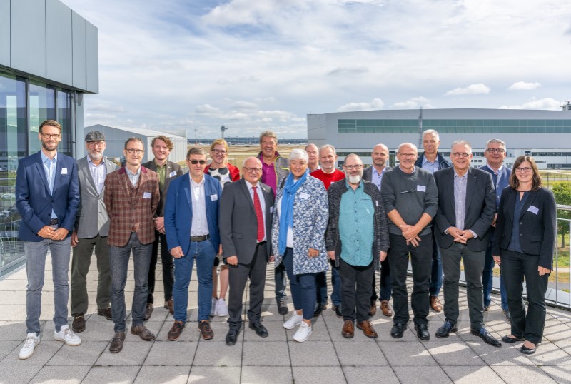 A large group of people pose smiling on a roof terrace against an airport background.