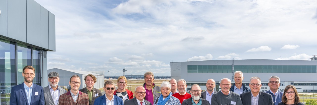 A large group of people pose smiling on a roof terrace against an airport background.