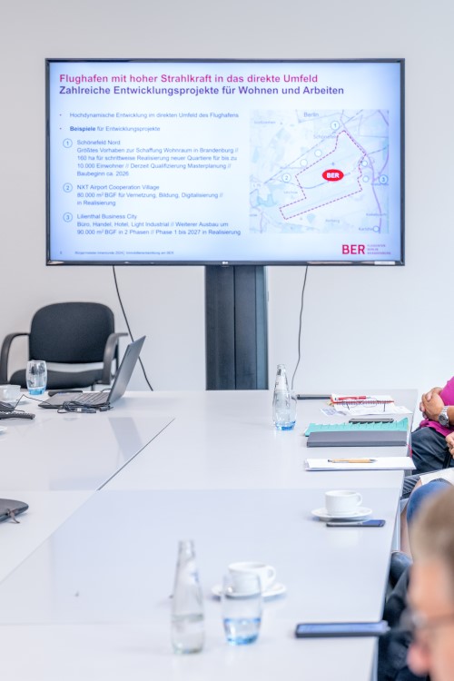 At the head of a conference table, at which people are sitting - only parts of which can be seen - there is a large screen showing a presentation on the importance of the airport for the surrounding area.