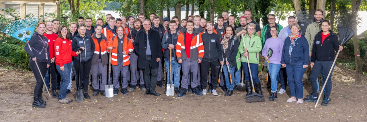 A large group is standing at the edge of a grove, posing for a photo. Some of them are holding gardening tools.