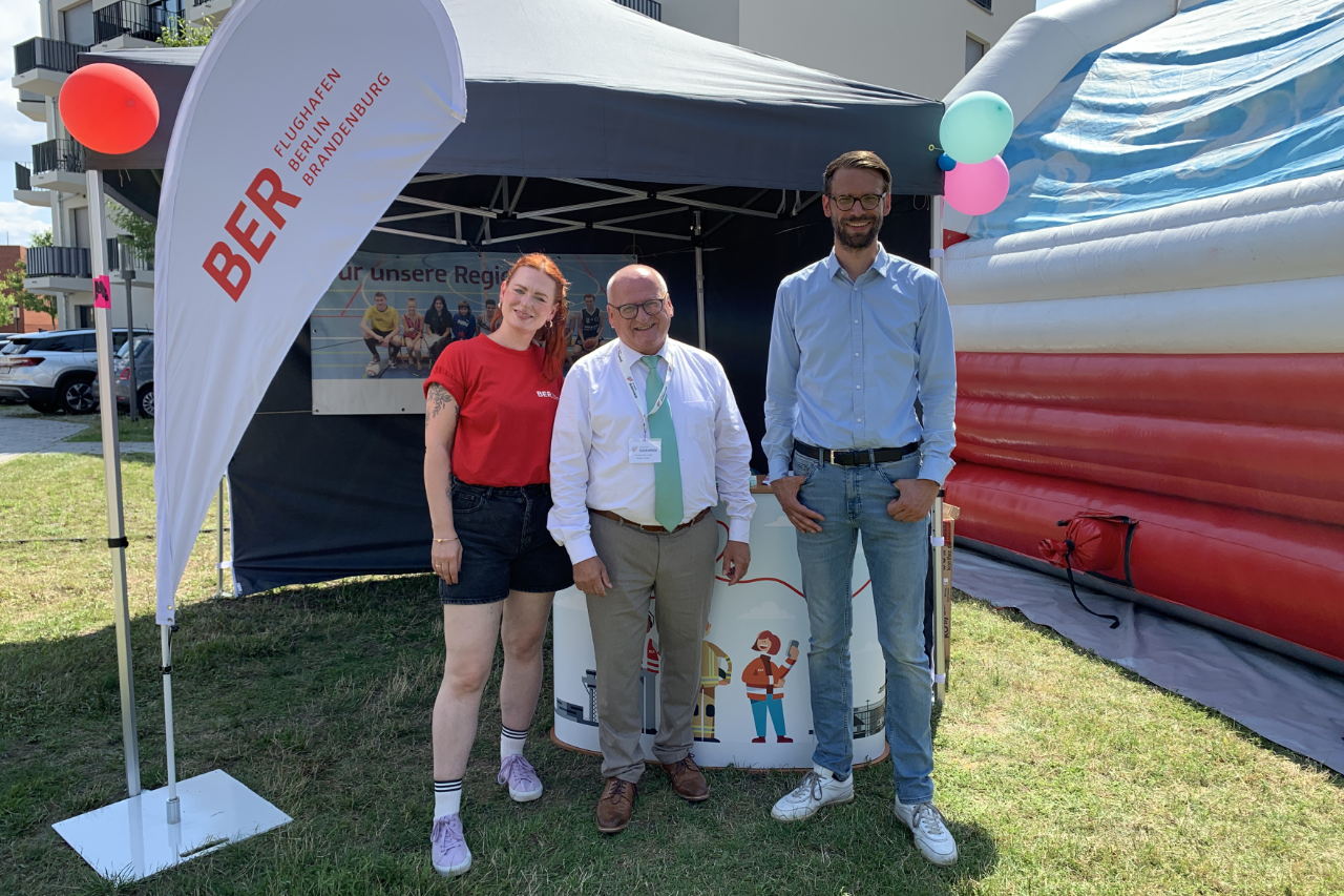 Three people stand in front of a BER information stand. In the background is a bouncy castle.