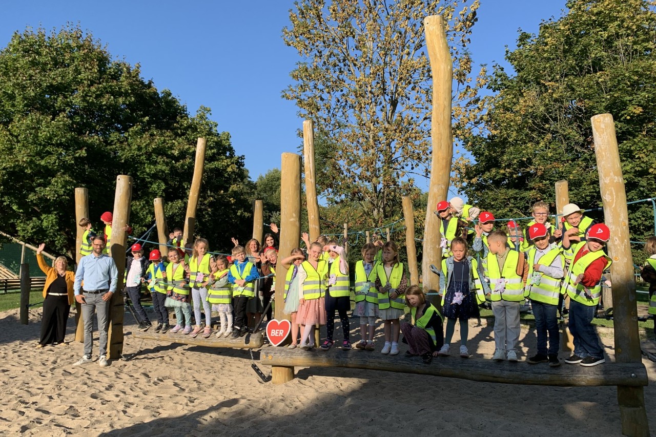 A large group of children pose in yellow high-visibility vests on a playground.