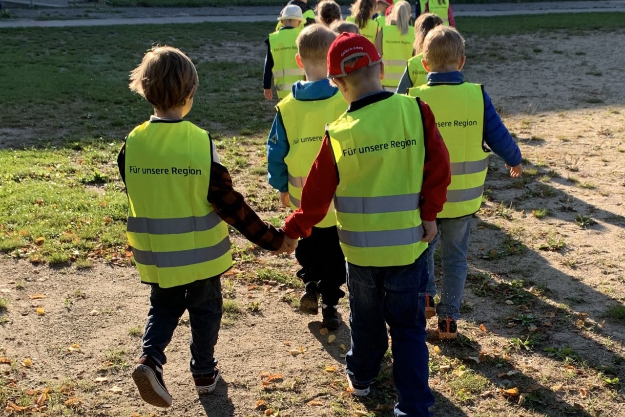 Children wearing yellow high-visibility vests are walking in rows of two with their backs to the camera.
