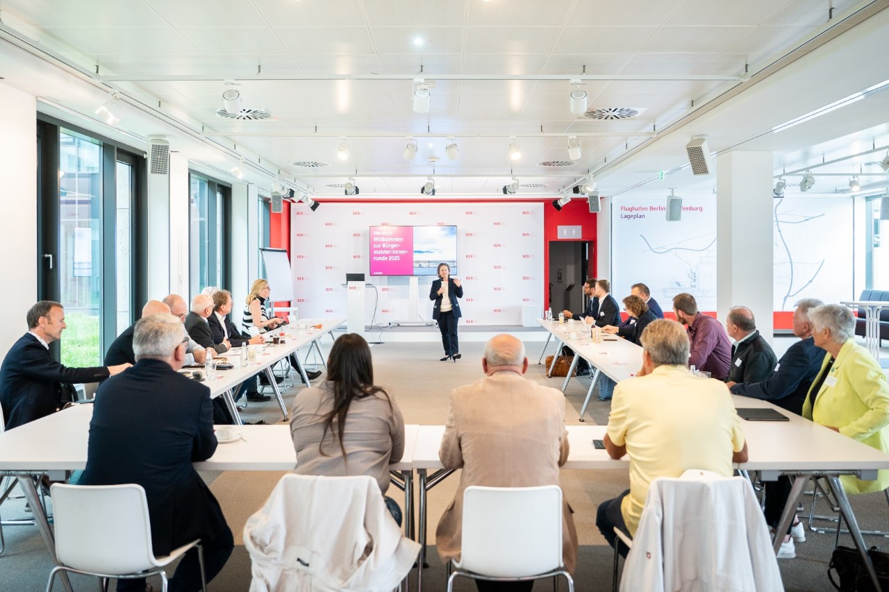 Many people sitting at tables arranged in a rectangle. At the front, one person is giving a presentation.