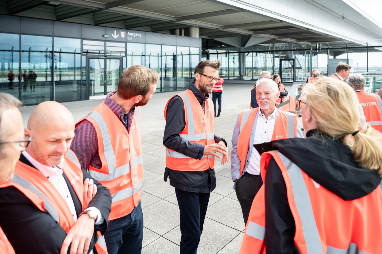 Several people wearing orange-red safety vests are standing on a terrace. One of them is talking to the others.