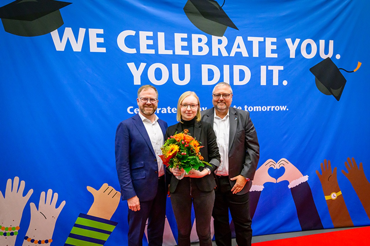 Three people pose smiling, the person in the middle is holding a bouquet of flowers.