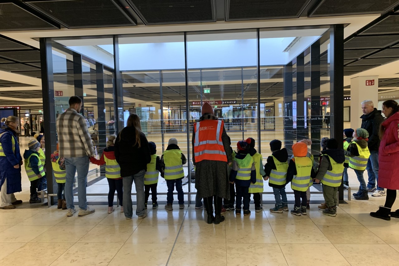 Children wearing yellow safety vests stand in front of a large glass front in a terminal.