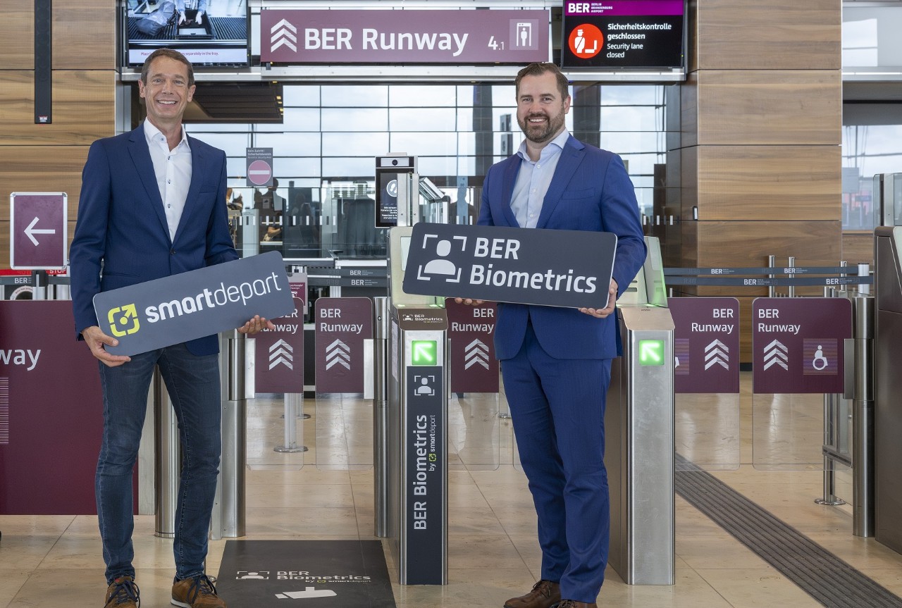 two people standing in front of BER Runway with signs in their hands