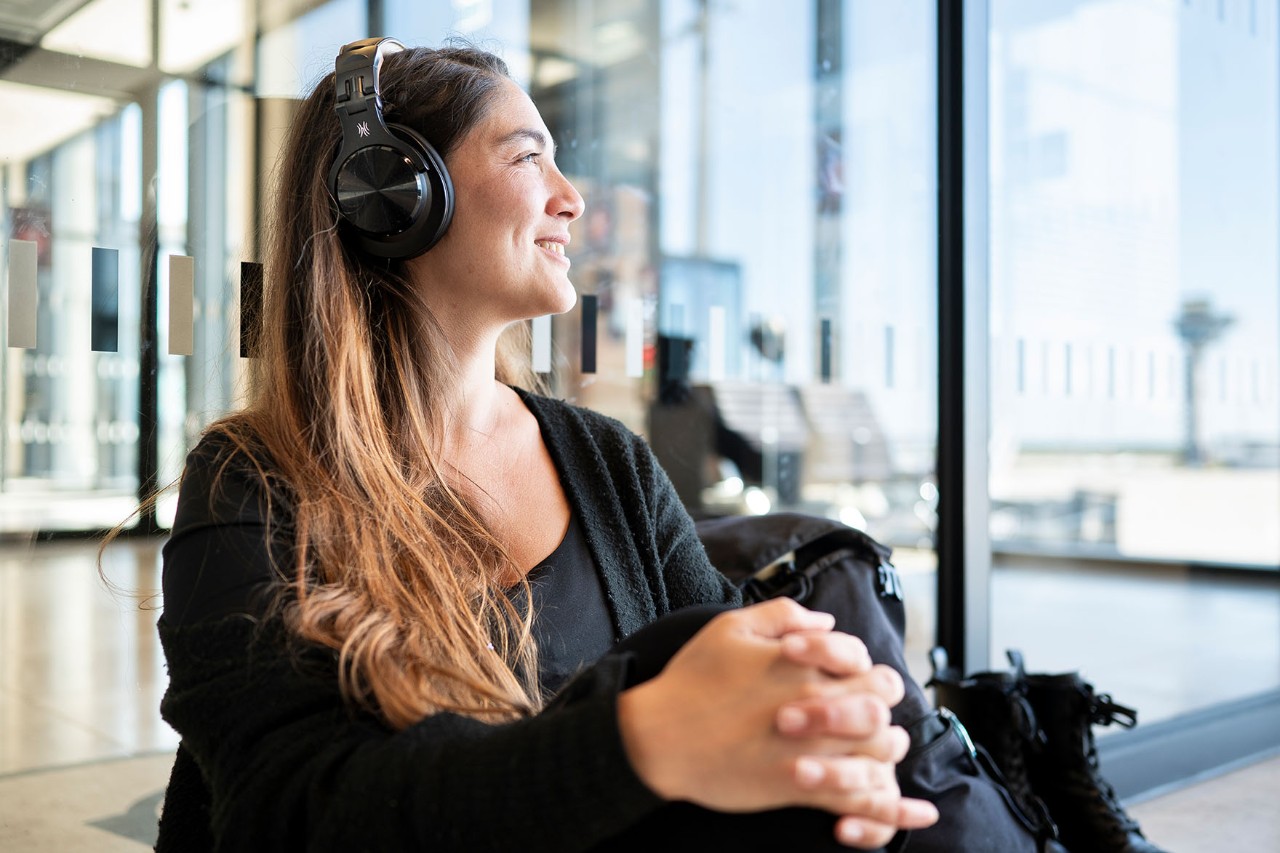 A traveler looks cheerfully out the window as she waits for her flight. She is wearing headphones. 