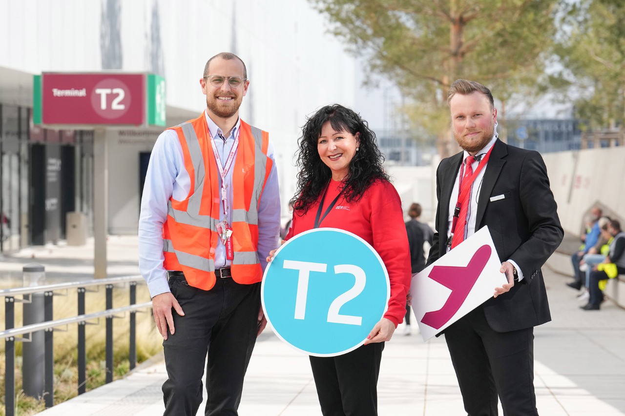 Three people are standing in front of Terminal 2 with colorful signs.